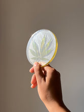 Load image into Gallery viewer, Pressed hemp leaf preserved in a glossy round resin coaster, held in hand for a natural and modern aesthetic.
