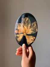 Load image into Gallery viewer, Tilted view of a black resin coaster highlighting a dried hibiscus flower with vivid petals and a distinct pollen stem. The natural light reflects off the glossy surface, enhancing the rich details and vibrant colors