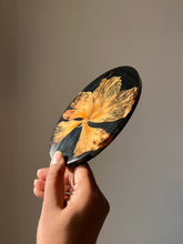 Load image into Gallery viewer, Tilted angle of an oval black resin coaster showcasing a large hibiscus flower with rich orange hues and a prominent pollen stem.