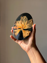 Load image into Gallery viewer, Top-down view of an oval black resin coaster featuring a vibrant dried hibiscus flower with intricate petals and a prominent pollen stem. The glossy finish enhances the flower’s texture, creating a striking contrast against the black background.