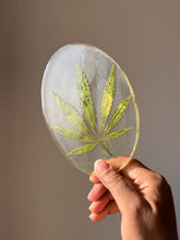 Load image into Gallery viewer, Hemp leaf resin coaster displayed at an angle, reflecting light to highlight its glossy finish and intricate details