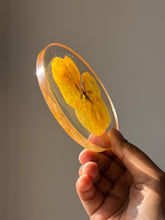Load image into Gallery viewer, Side angle of a resin coaster with a full yellow pansy flower embedded in clear resin. The flower’s bright yellow petals cover the entire coaster, with the smooth edges and glossy finish enhancing the flower’s vivid colors
