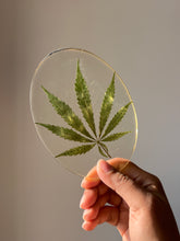 Load image into Gallery viewer, Flat lay of an oval resin coaster with a green hemp leaf design, styled with minimalistic decor elements

