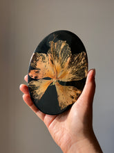 Load image into Gallery viewer, Hand holding an oval black resin coaster with a preserved hibiscus flower. The vivid petals and pollen stem stand out vividly against the sleek black background, highlighted by soft natural light.
